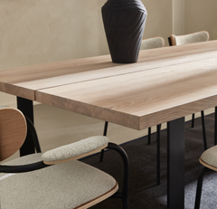 Nærbillede af et spisebord i lys eg med sorte ben, beige polstrede stole og en mørk vase.
