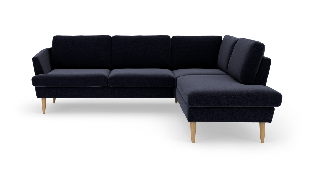 Mørkeblå L‑sofa med chaiselong til højre og lyse træben, frilagt på hvid baggrund.