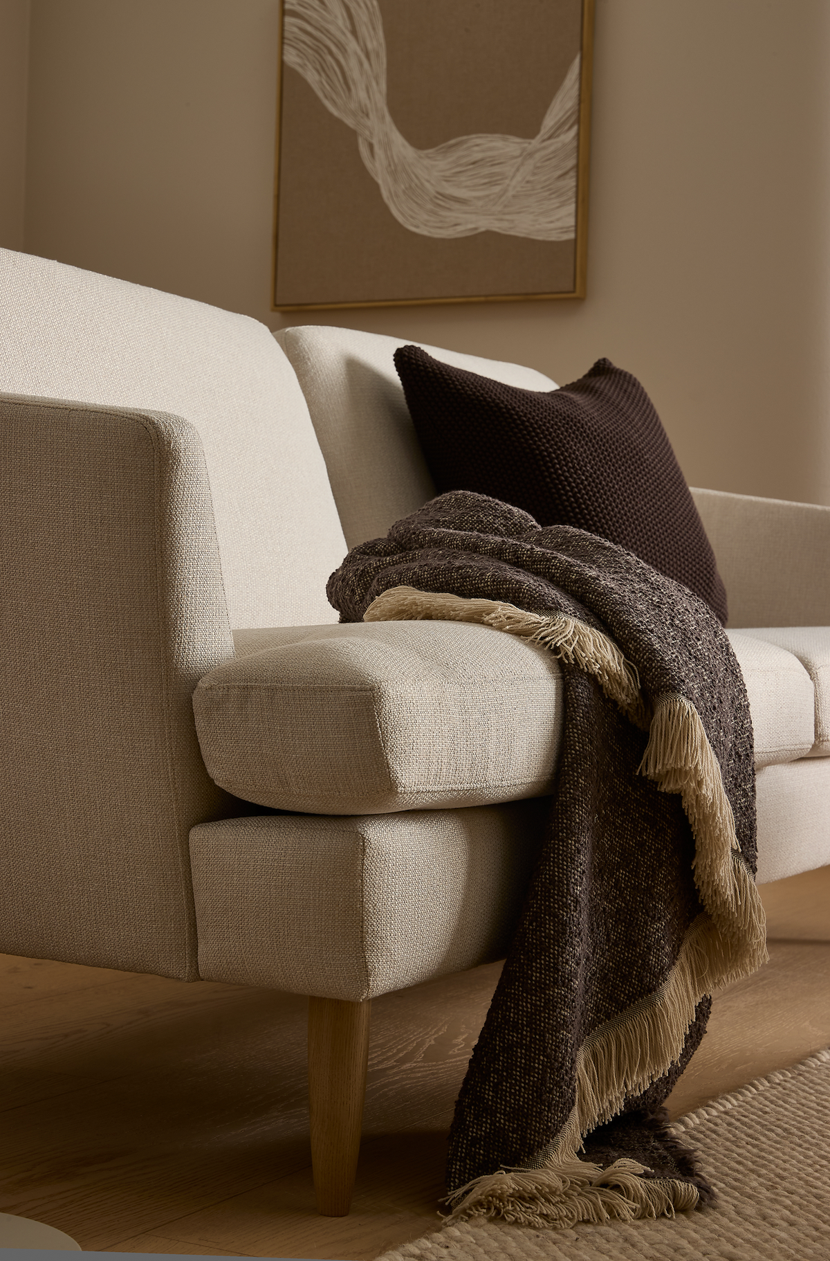 Beige sofa med mørkebrun pude og brun plaid med frynser over armlænet, træben, neutral måtte og et abstrakt vægbillede.