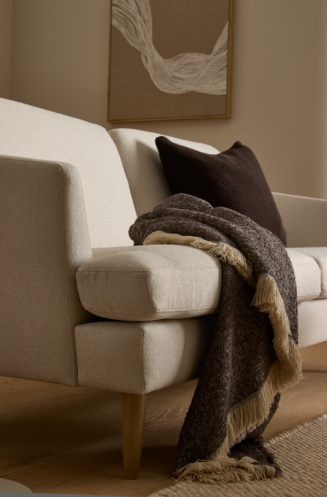 Beige sofa med mørkebrun pude og brun plaid med frynser over armlænet, træben, neutral måtte og et abstrakt vægbillede.