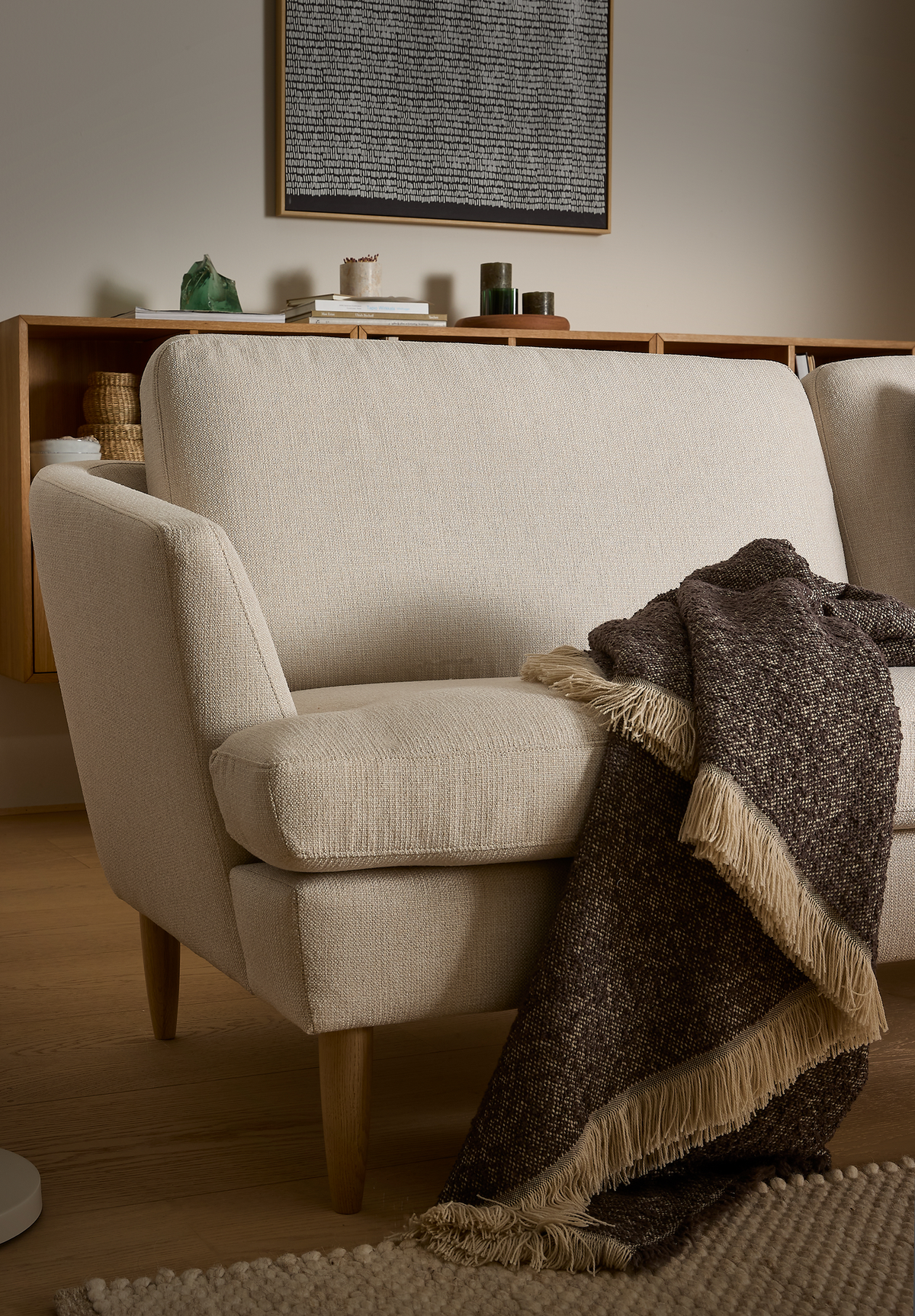 Beige to-personers sofa med træben og en brun plaid med frynser over armlænet; i baggrunden en skænk med bøger, lys og kurve samt et abstrakt vægbillede.