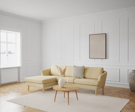 Lyst stue med gul hjørnesofa, sofabord i lyst træ, neutralt tæppe og minimalistisk vægkunst på panelvæg; dagslys fra vindue.