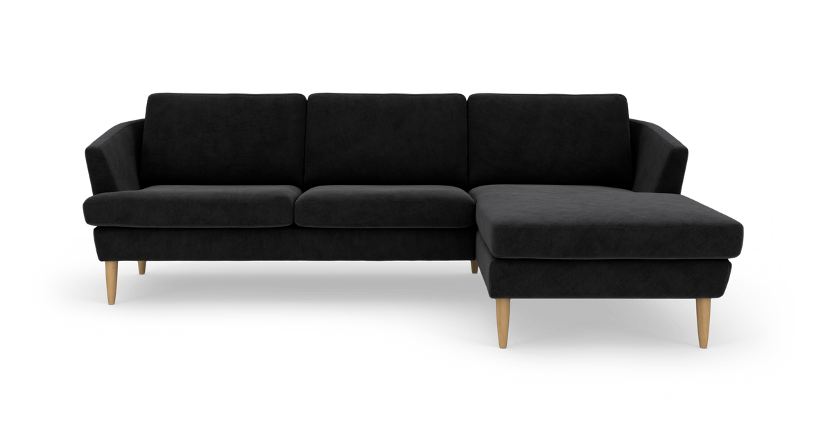 Sort hjørnesofa med chaiselong til højre og lyse træben, frilagt på hvid baggrund.