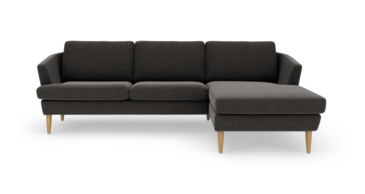 Mørkegrå sofa med chaiselong til højre og lyse træben, frilagt på hvid baggrund.