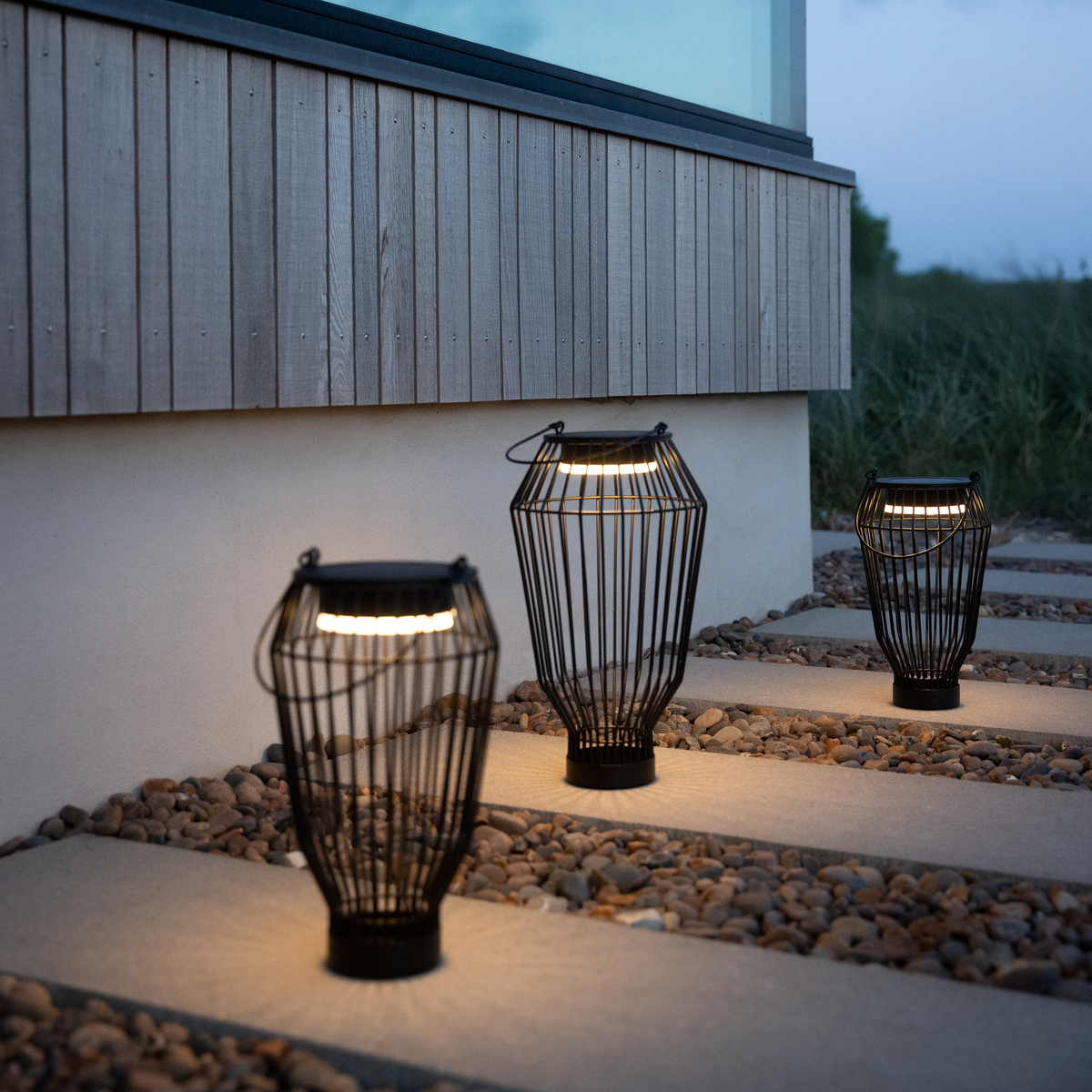 Tre sorte LED-lanterner oplyser en stenbelagt sti ved et moderne hus i skumringen.
