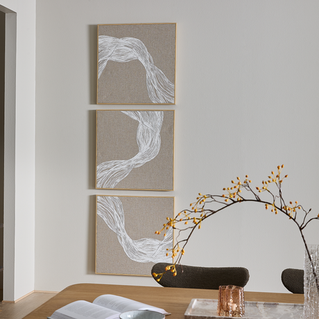 Tre beige vægbilleder med hvid abstrakt linjekunst over spisebord med vase og gren samt fyrfadslys, i dagslys.