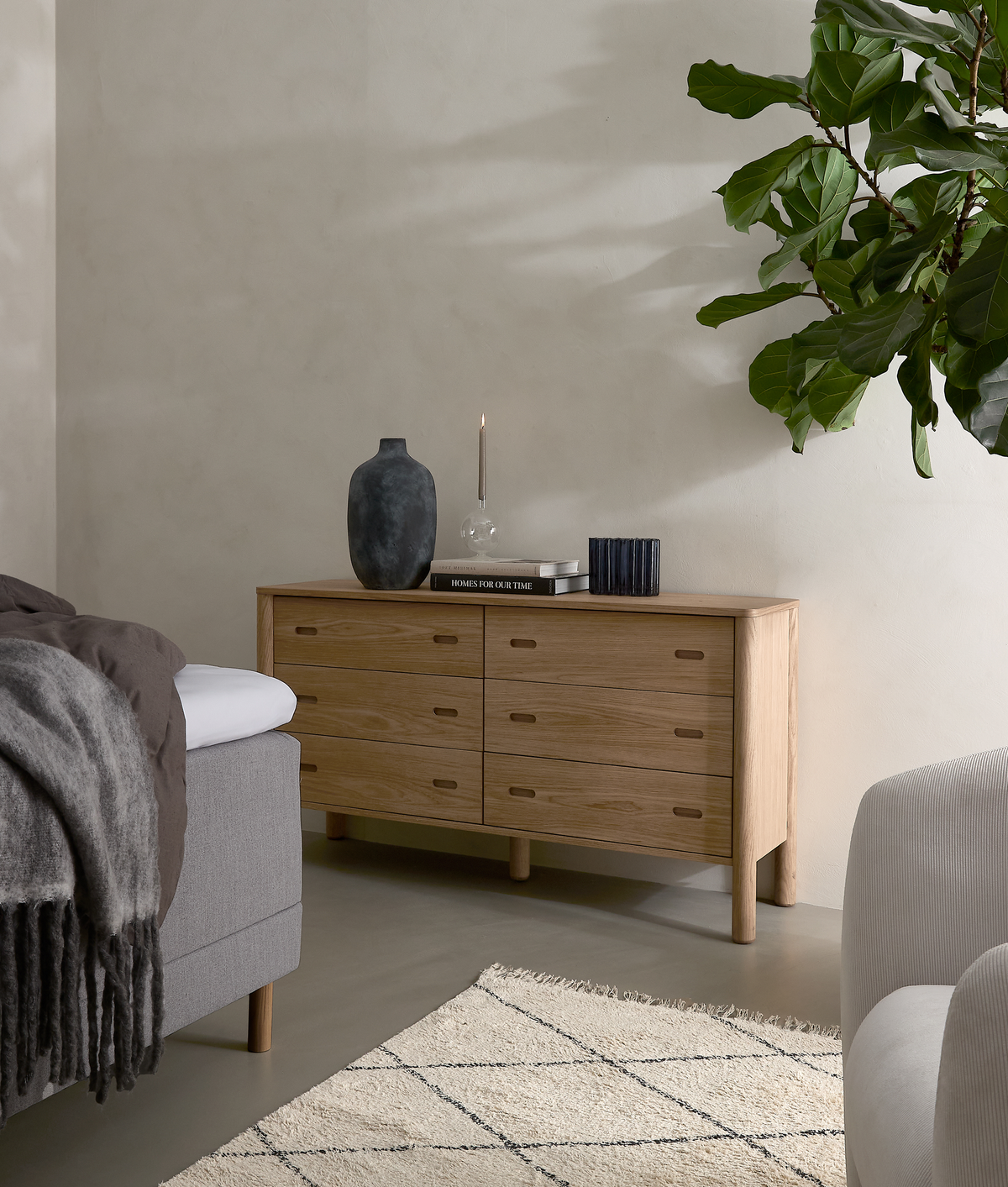 Lyst, minimalistisk soveværelsesmiljø med egetræskommode med seks skuffer, pyntet med mørk vase, slank lysestage og bøger, hvoraf en har titlen ”HOMES FOR OUR TIME”; stor grøn stueplante, seng med plaid og lys tæppe.