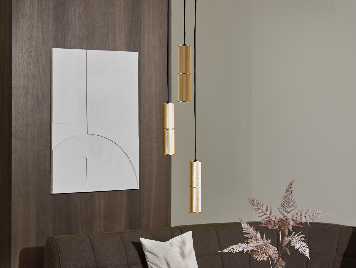 Moderne stue med mørkebrun sofa, hvid pude, tørrede kviste i en vase, abstrakt hvid vægkunst og tre cylindriske messingpendler i forskellige højder.