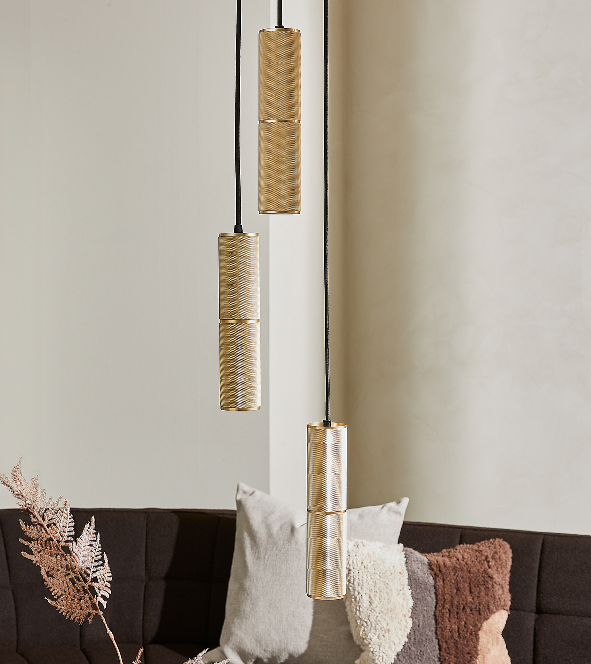 Tre slanke cylindriske pendellamper i messinglook hænger over en mørk sofa med teksturerede, neutrale puder i en minimalistisk stue.