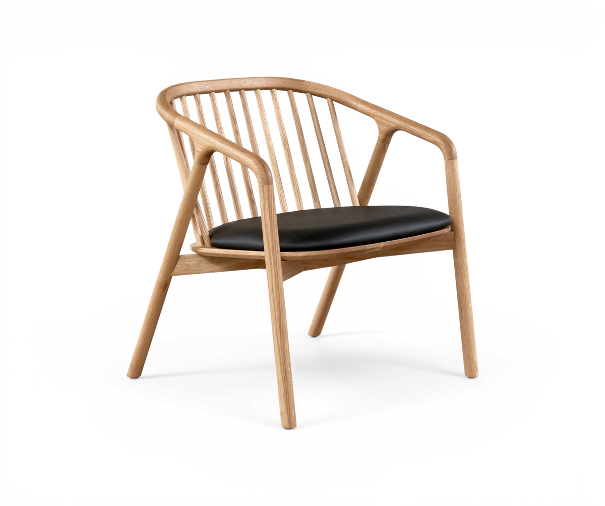 Stol i lyst træ med afrundede armlæn og spindestænger i ryggen, sort polstret sæde – moderne skandinavisk design i studiemiljø.