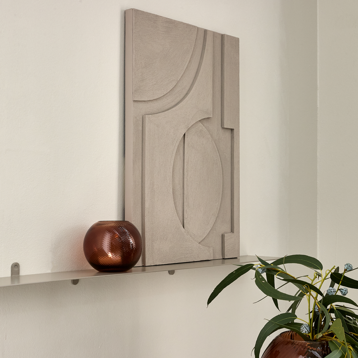 Abstrakt relief vægkunst i beige står på en smal hylde sammen med en brun glasvase og en grøn plante.