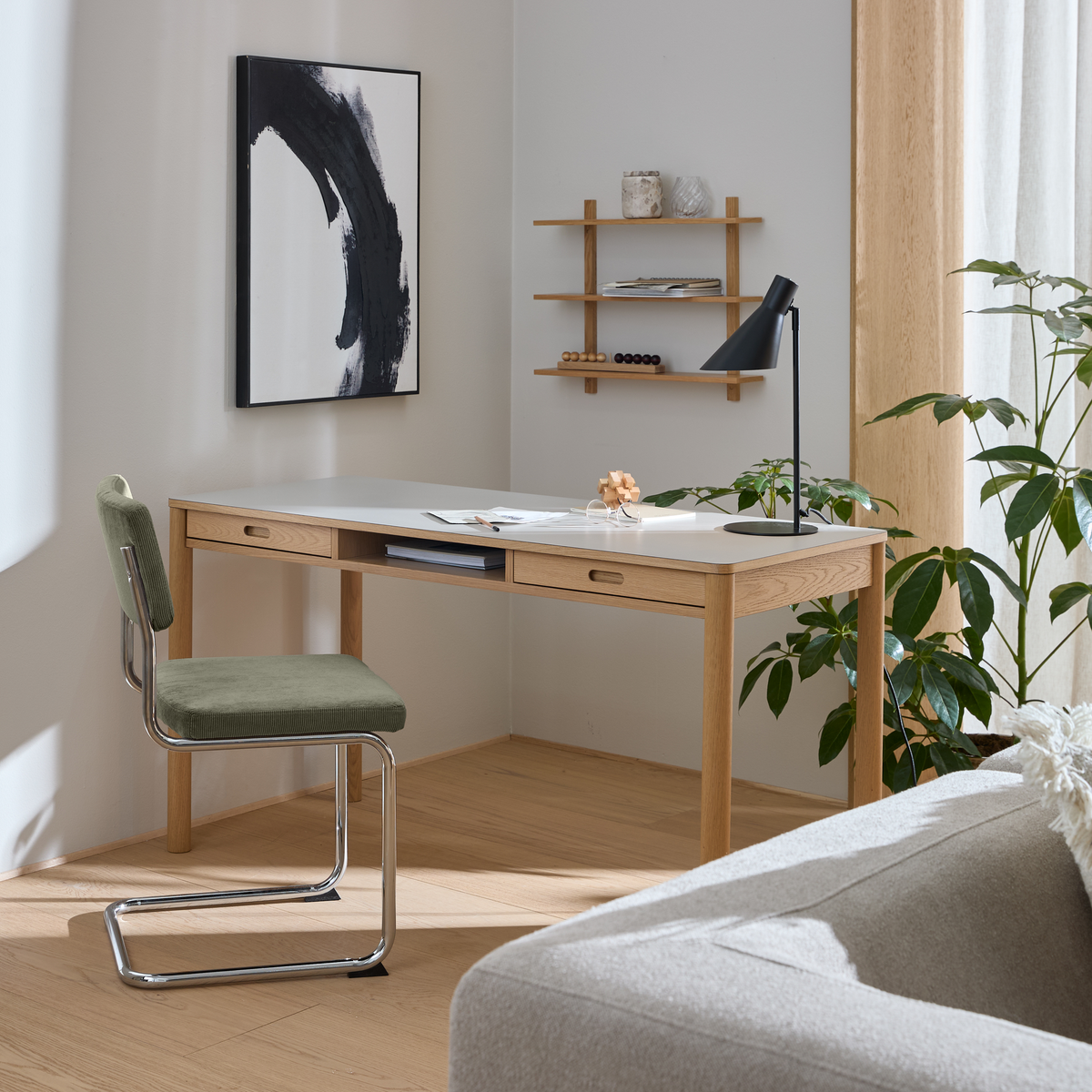 Lyst hjemmekontor med træskrivebord, grøn stol, sort bordlampe, vægkunst og grønne planter ved vindue.