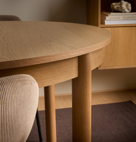 Nærbillede af et afrundet spisebord i lyst egetræ med cylindriske ben og en beige riflede stol; i baggrunden en hylde med bøger – KINFOLK, WILDERNESS – og en dekorativ sten i et varmt, minimalistisk interiør.