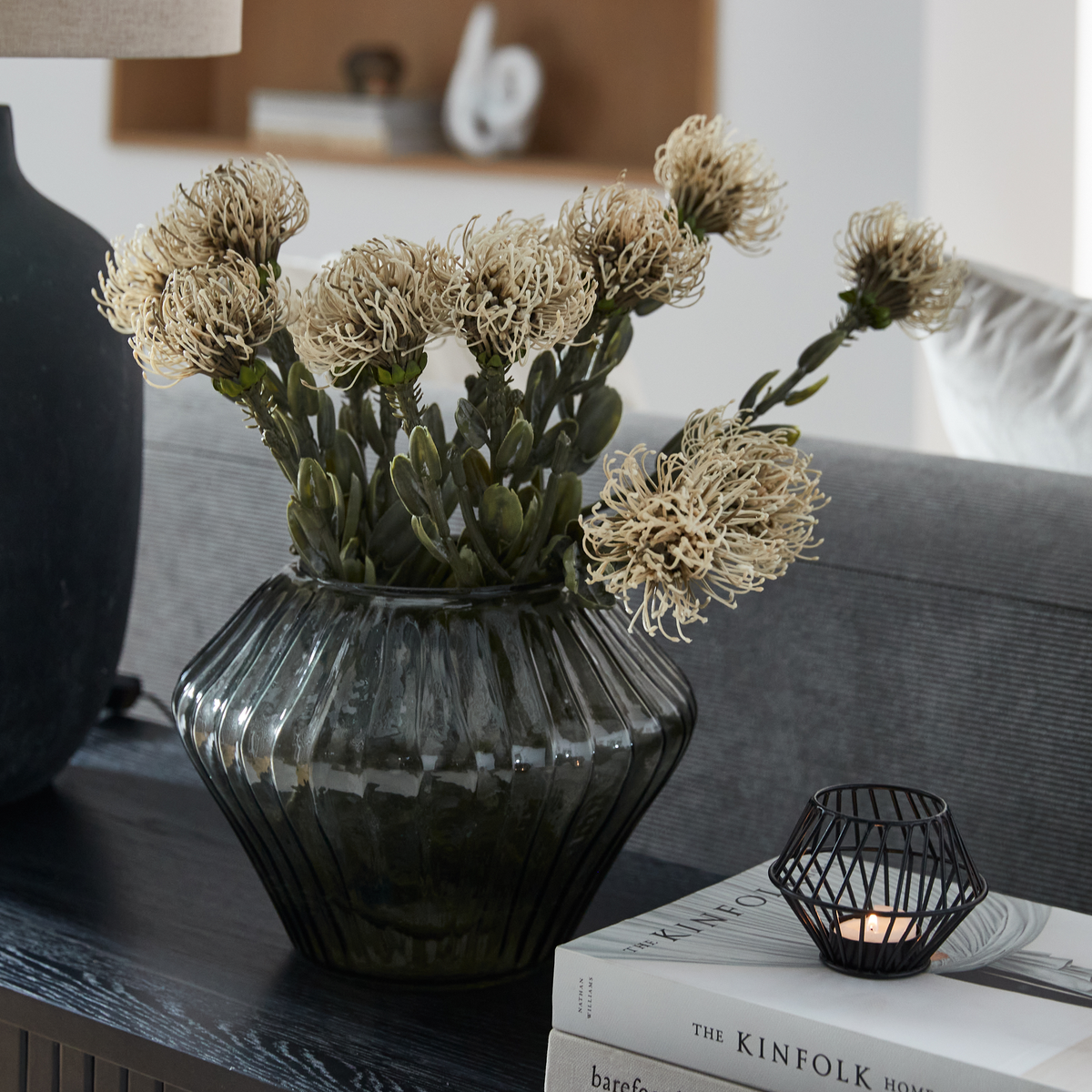 Mørk riflet glasvase med lyse tørrede blomster på sort bord, ved siden af tændt fyrfadsstage og bogen The Kinfolk Home.