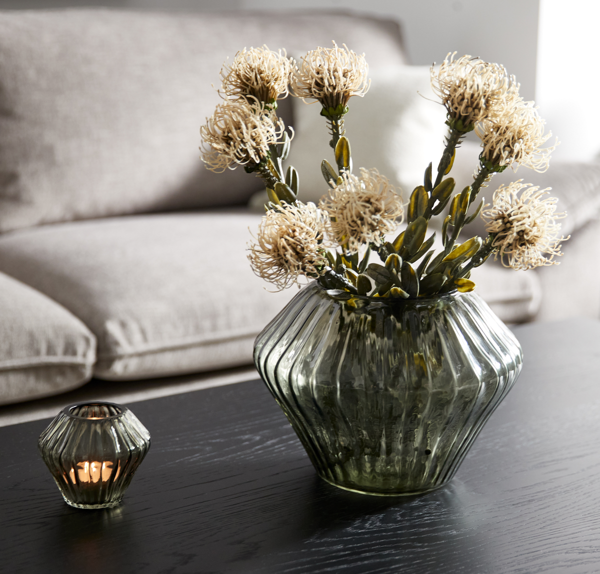 Grøn riflet glasvase med lyse blomster på sort sofabord ved siden af en matchende fyrfadsstage, beige sofa i baggrunden.