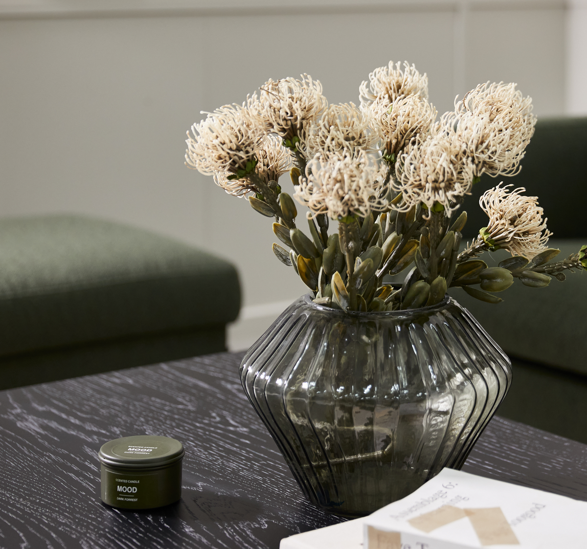Glasvase med tørrede blomster på mørkt sofabord ved siden af en lille grøn dåse med teksten ”MOOD Scented Candle – Green Forest” i en stue med grøn sofa.