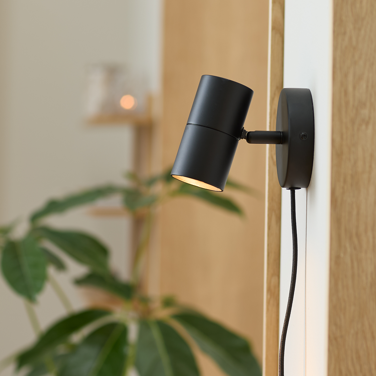 Sort vægmonteret spot med ledning, tændt, på hvid væg ved træpanel med grøn plante sløret i baggrunden.