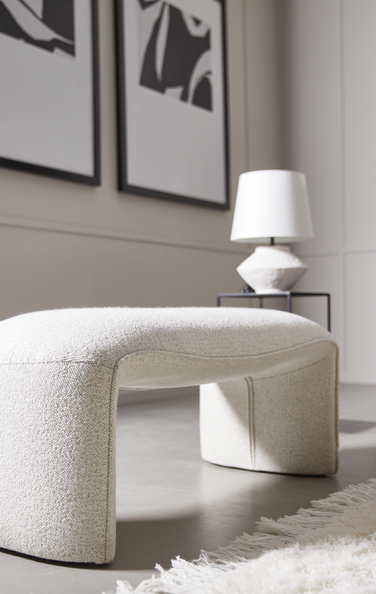 Lys beige bouclébænk i moderne interiør med skulpturel bordlampe, sort-hvid abstrakt vægkunst og hvidt ryetæppe med frynser.