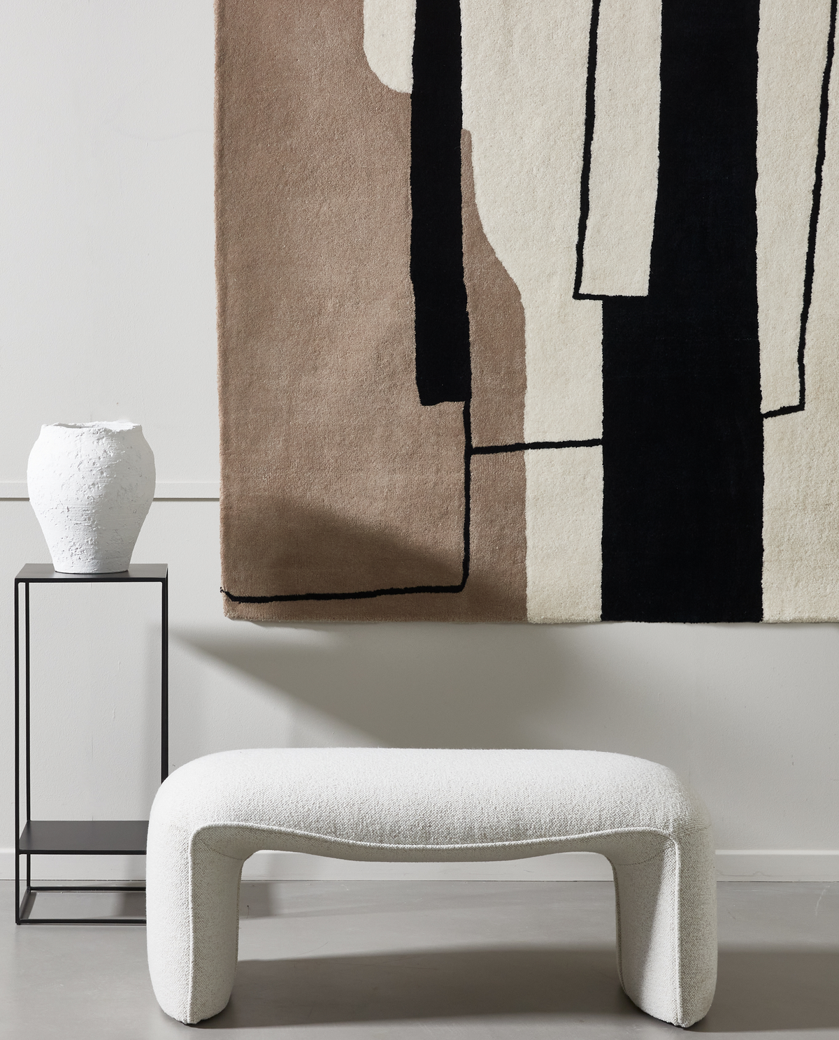 Minimalistisk indretning med hvid bouclébænk, sort metalbord med en hvid keramisk vase og abstrakt vægtæppe i beige, hvid og sort.