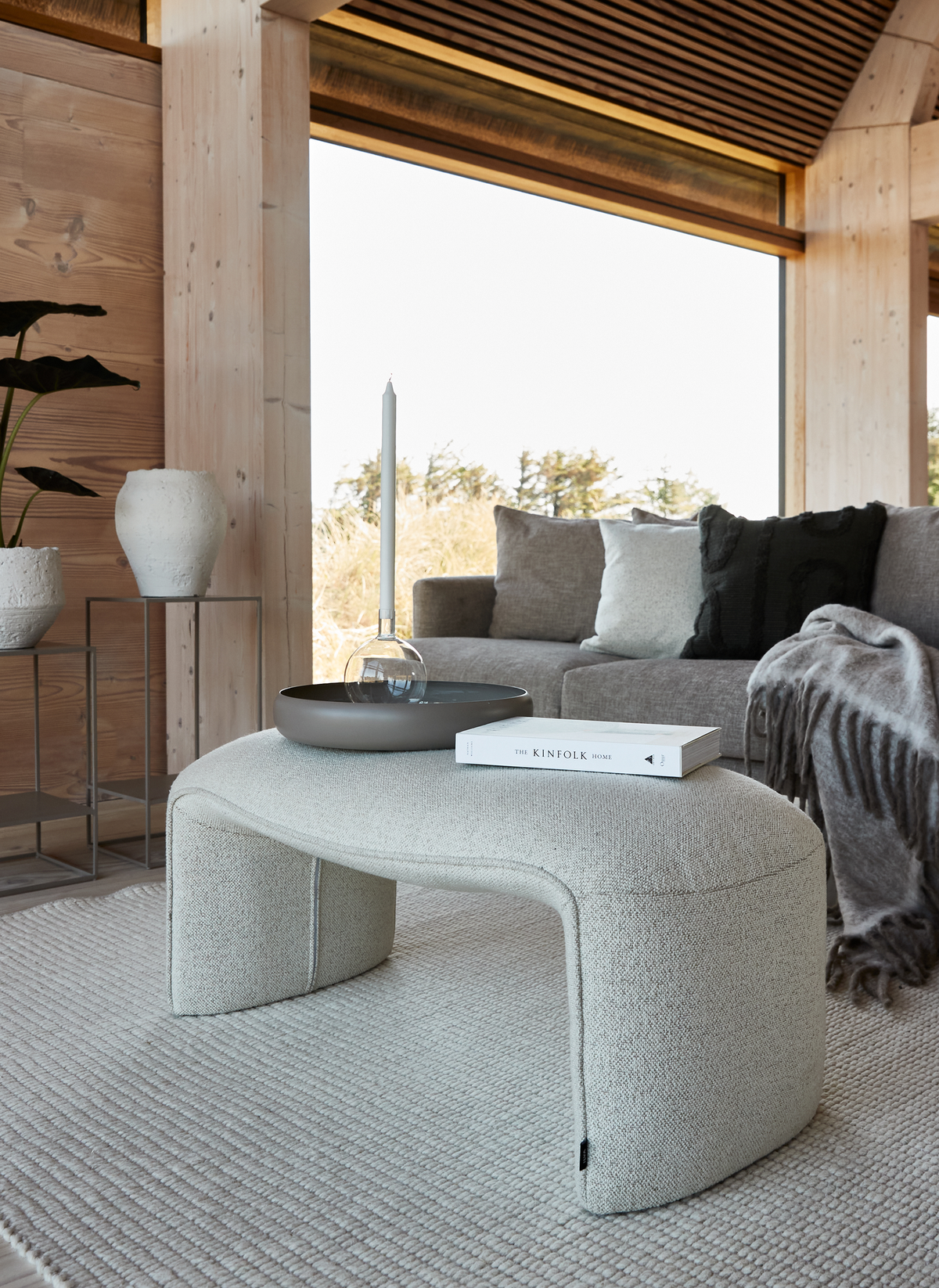 Lys stue i neutrale toner med buet bouclé-ottoman som sofabord, en rund bakke med glaslysestage med slankt lys og bogen The Kinfolk Home; grå sofa med puder og plaid, store vinduer og lyse trævægge.
