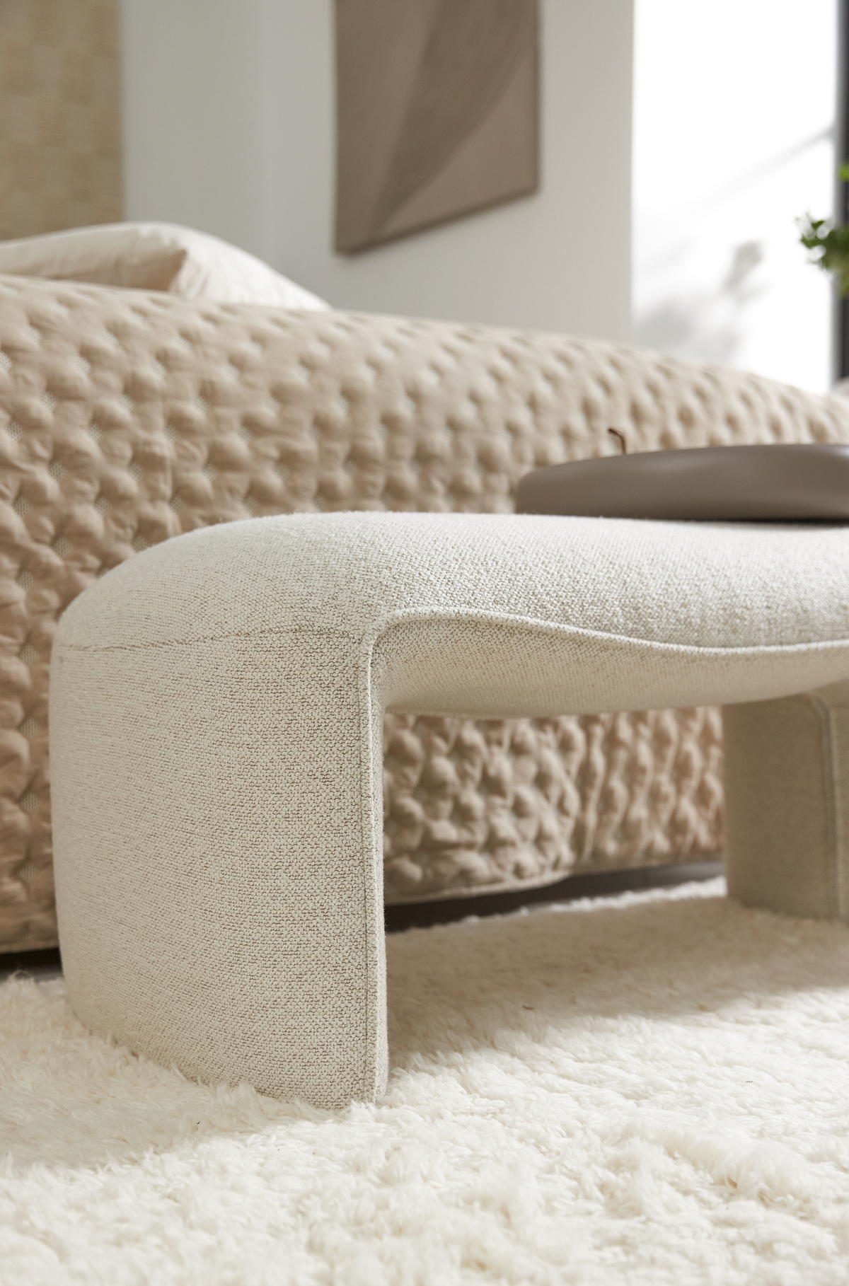Nærbillede af en cremefarvet bouclébetrukket, buet bænk for enden af en seng med et beige, tekstureret sengetæppe og et blødt hvidt ryatæppe i et lyst, minimalistisk soveværelse.