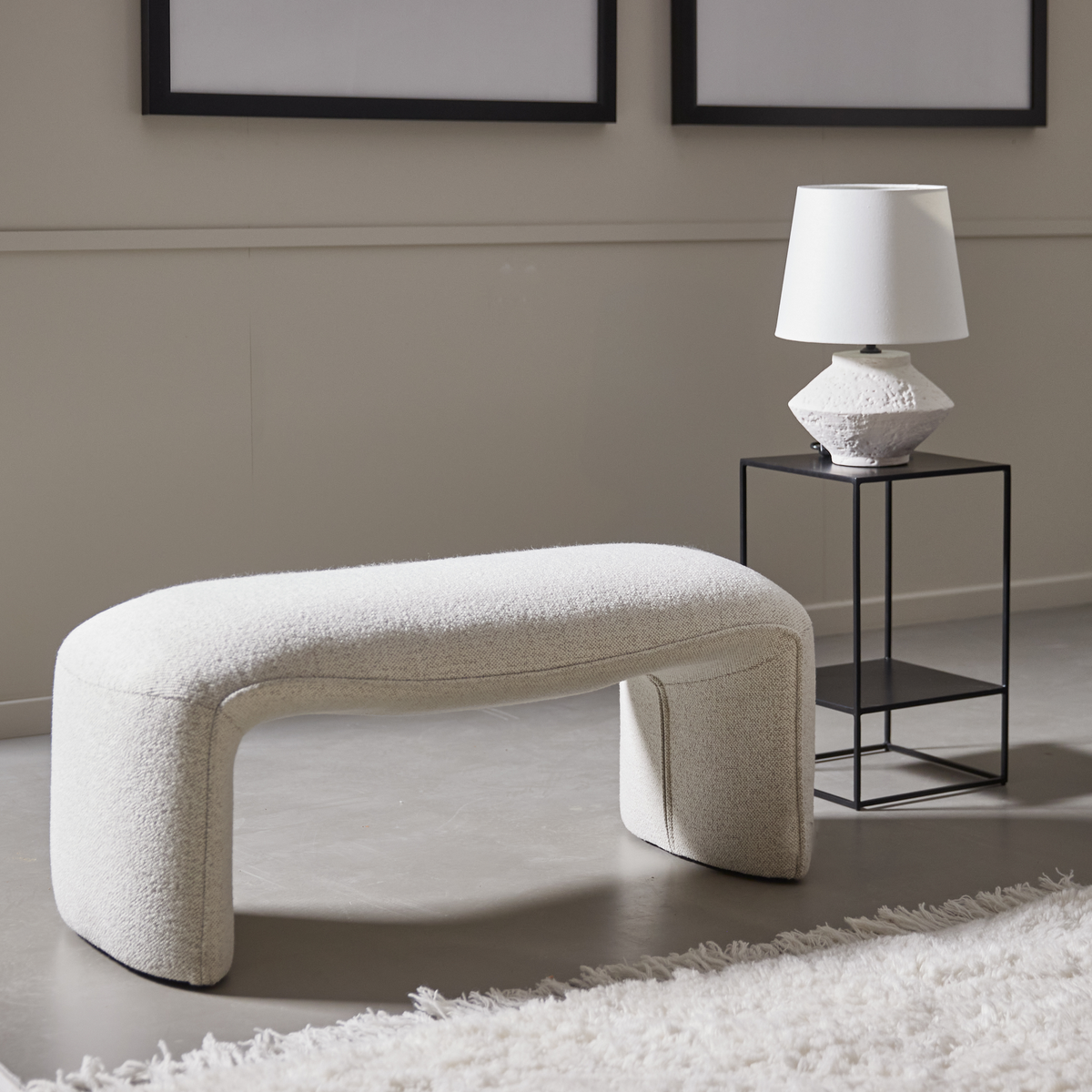 Lys bouclé-bænk med bløde former ved siden af et sort sidebord med hvid bordlampe i et minimalistisk stuemiljø.