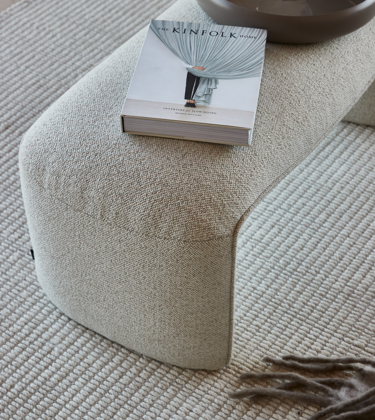 Bogen The Kinfolk Home – Interiors for Slow Living af Nathan Williams ligger på en lys, tekstureret skammel på et beige, vævet uldtæppe med et blødt plaid i kanten.