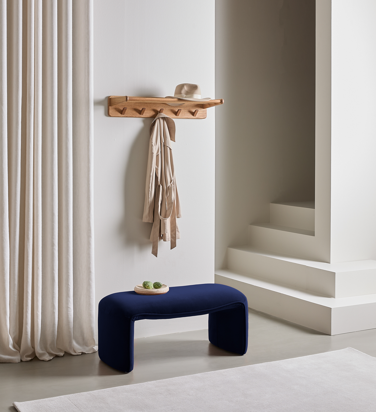 Minimal entré med vægmonteret knagerække i træ med beige frakke og hat, mørkeblå bænk og lyse gardiner ved hvide trin.