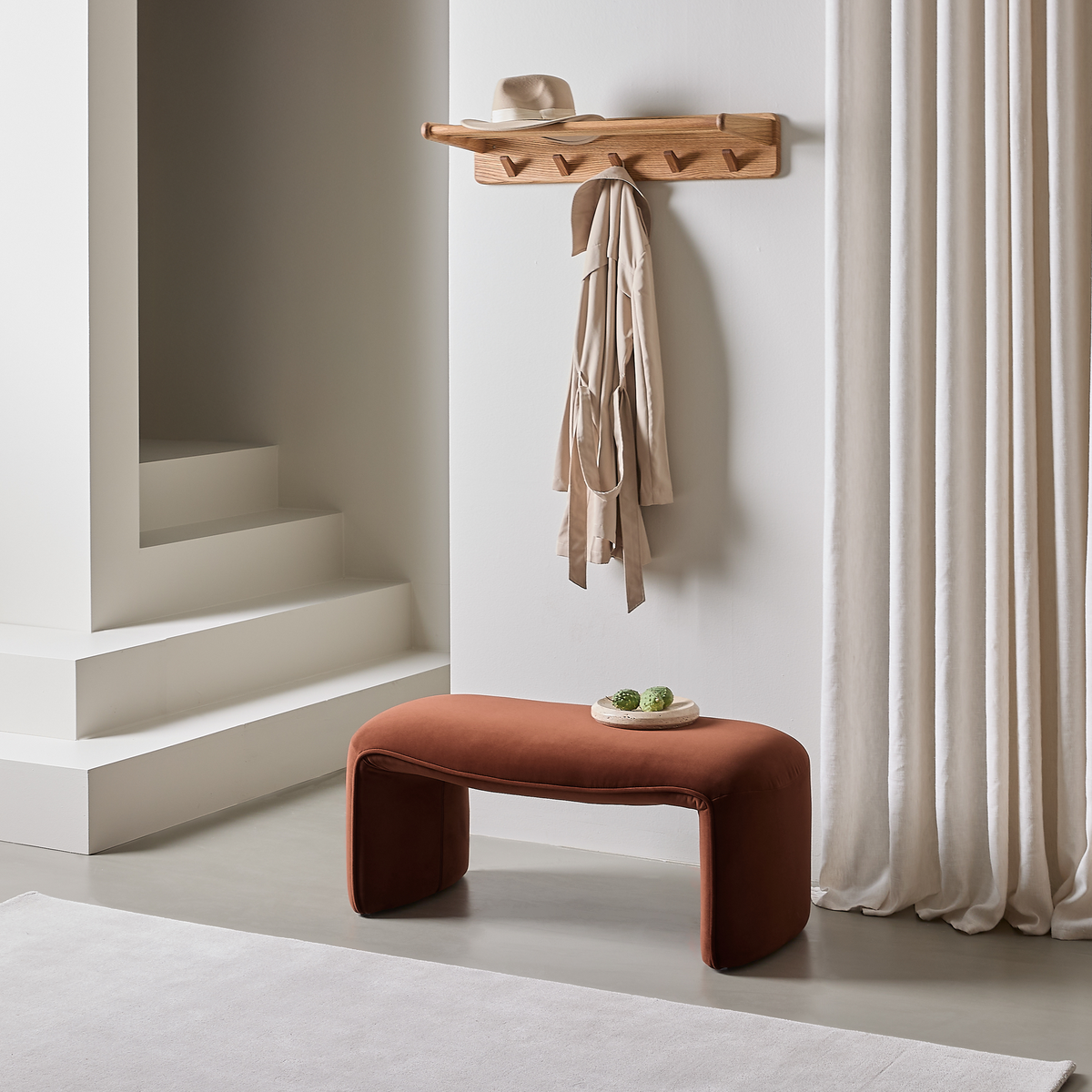 Minimal entré med terrakottafarvet bænk, træknagerække med beige frakke og hat, hvide gardiner og lys tæppe ved trappe.