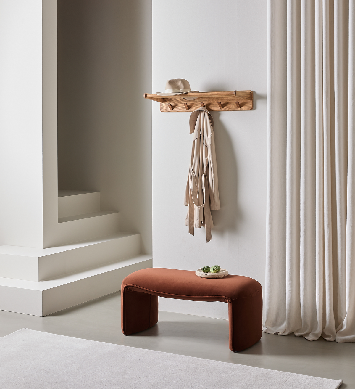 Minimalistisk entré med knagerække i træ med hat og beige frakke, terrakottafarvet bænk og lyse gardiner.