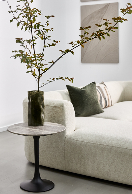 Lys, minimalistisk stue med lys buet sofa, grøn pude og en gren i mørkegrøn vase på et lille rundt sidebord med stenplade; neutral vægkunst i baggrunden.