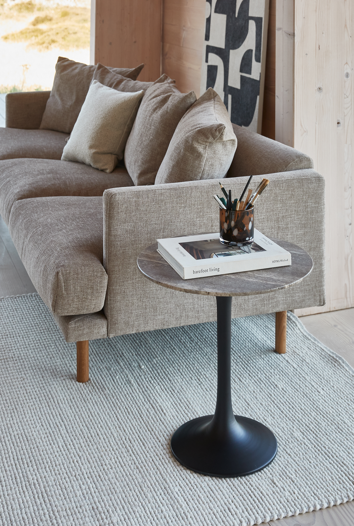 Beige sofa med rundt sidebord i sten og sort sokkel, bogen ”barefoot living” og blyanter i kop på et lyst tæppe i en skandinavisk stue.