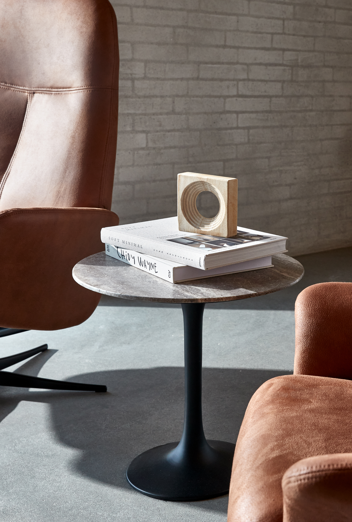 Rundt sidebord med sort tulipanfod og stentop, to bøger – bl.a. Soft Minimal – med en lille træskulptur ovenpå; brune læderstole og murstensvæg i blødt dagslys.