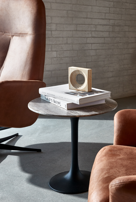 Rundt sidebord med sort tulipanfod og stentop, to bøger – bl.a. Soft Minimal – med en lille træskulptur ovenpå; brune læderstole og murstensvæg i blødt dagslys.