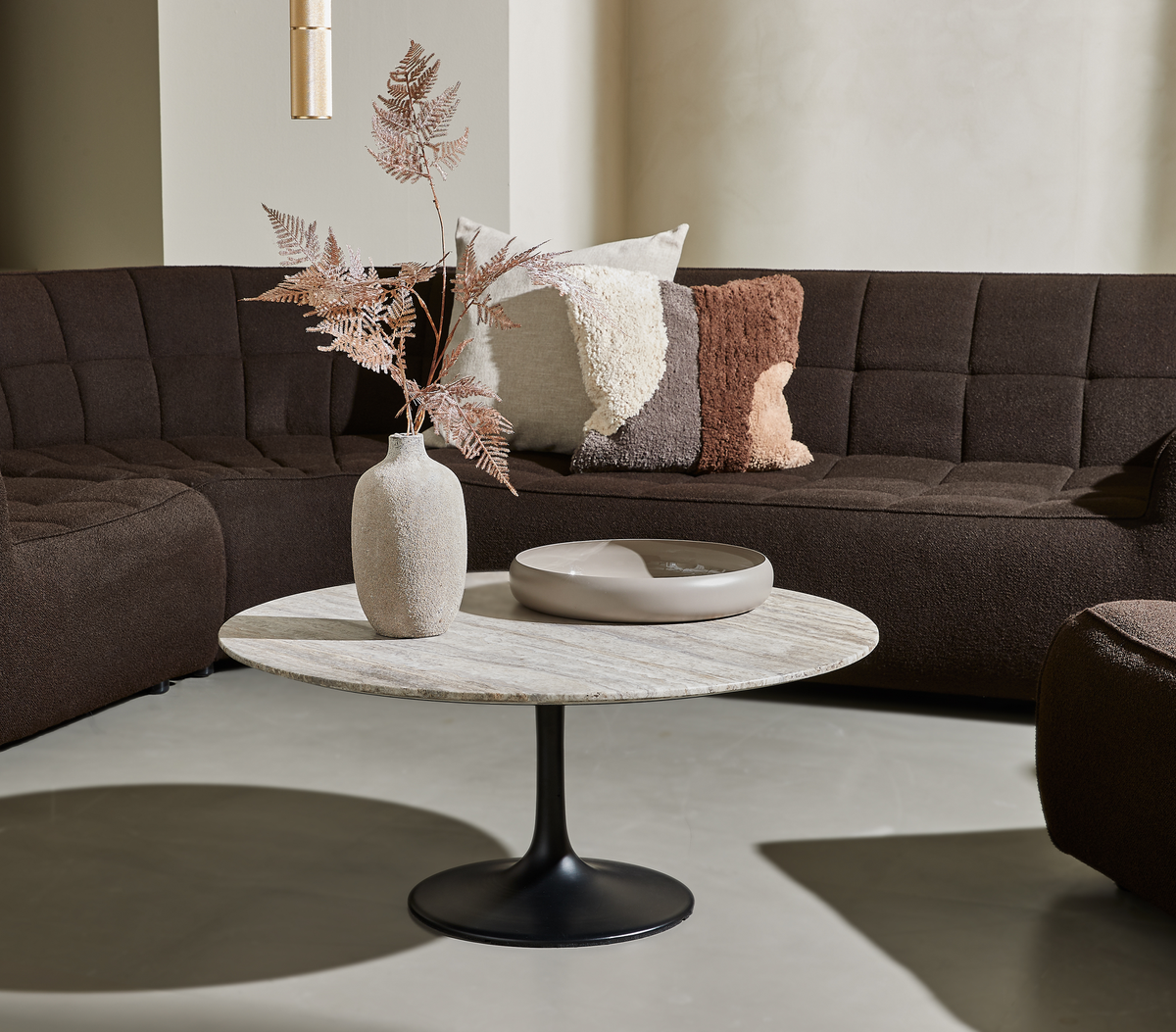 Moderne stue med mørk modulsofa, rundt sofabord med stenplade og sort fod, beige vase med tørrede grene og puder i brune nuancer.