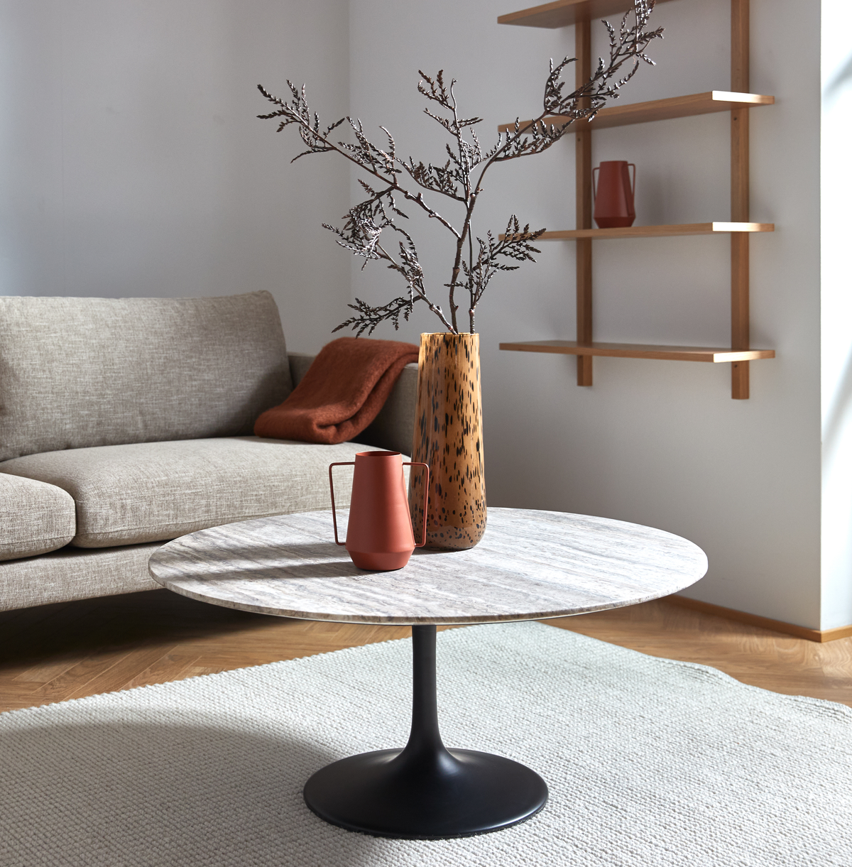 Lyst stue med rundt stenbord på sort piedestal, vase med grene og terrakotta-kande, beige sofa, hylder og lyst tæppe.