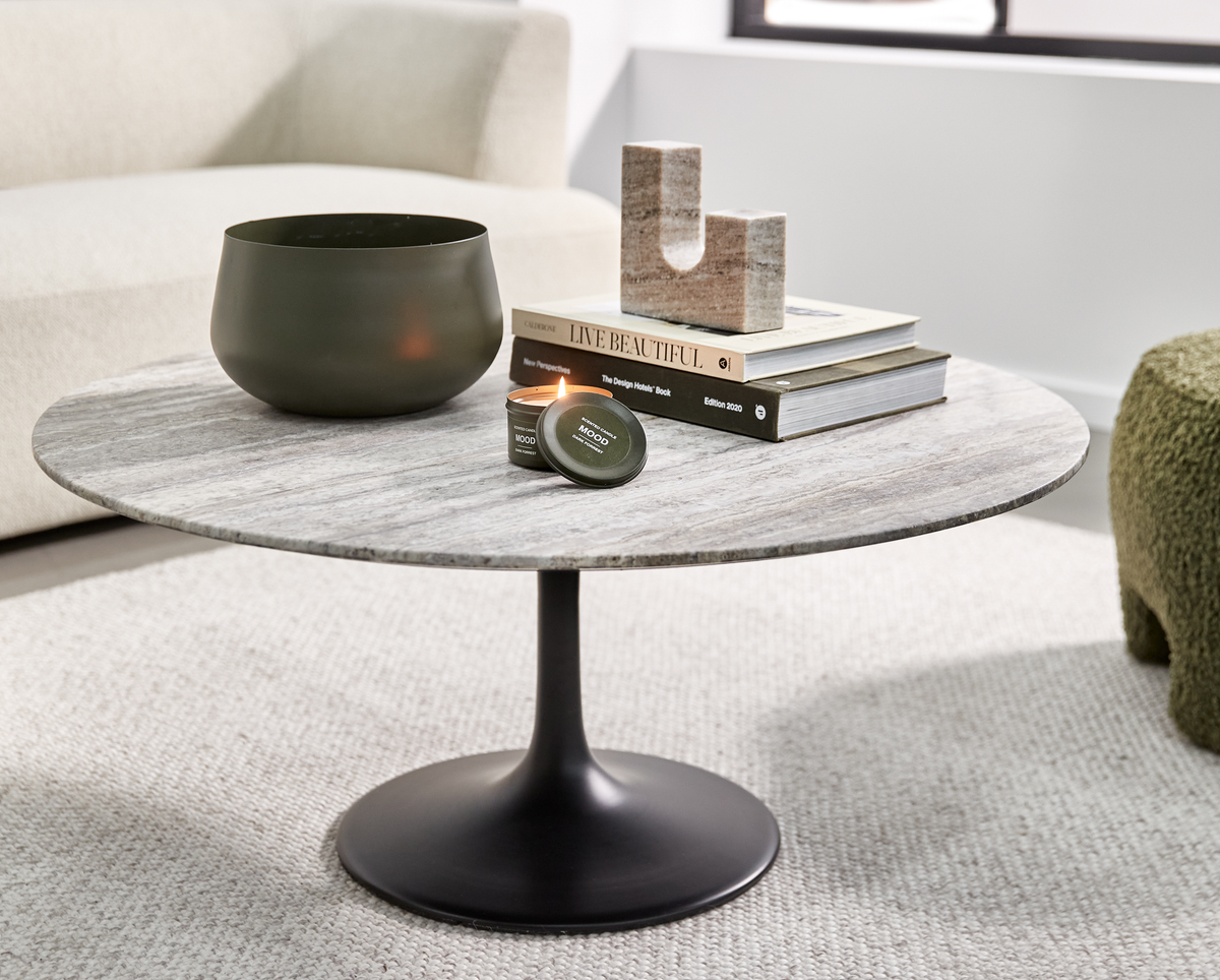 Rund sofabord med stenplade og sort søjlefod; grøn skål, tændt lille lysdåse mærket ”MOOD”, samt bogstak med ”Live Beautiful” og ”The Design Hotels Book Edition 2020” og en U‑formet stenskulptur; lyst stue-miljø.