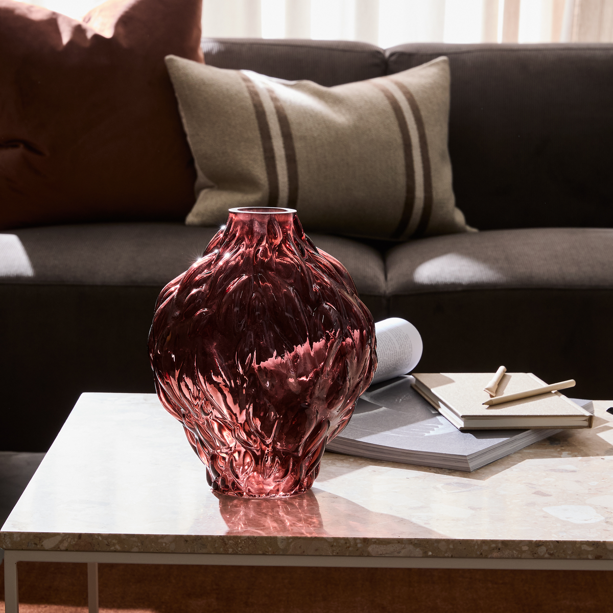 Vinrød skulpturel glasvase på sofabord foran mørk sofa med puder; notesbøger og magasin ved siden af.