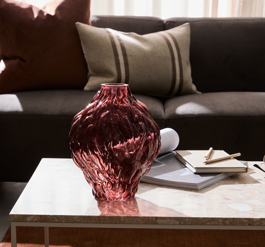 Rød, struktureret glasvase på et marmorbord i en stue med mørk sofa og beige puder, ved siden af notesbøger og blyant.
