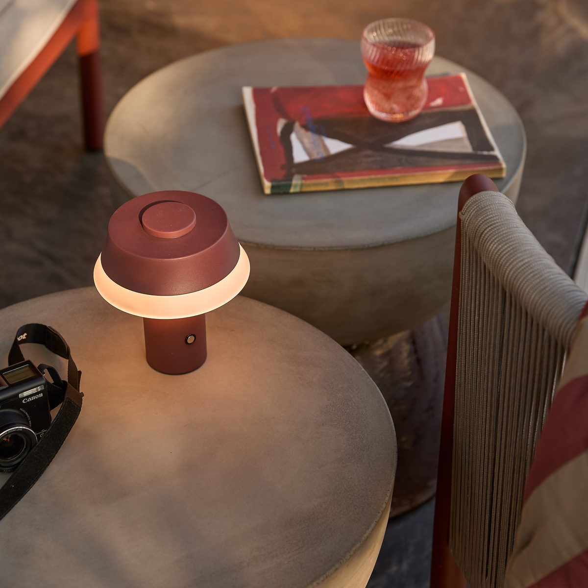 Bærbar bordlampe i bordeaux, tændt på runde betonborde; ved siden af ligger en bog med abstrakt omslag, et glas og et Canon‑kamera.