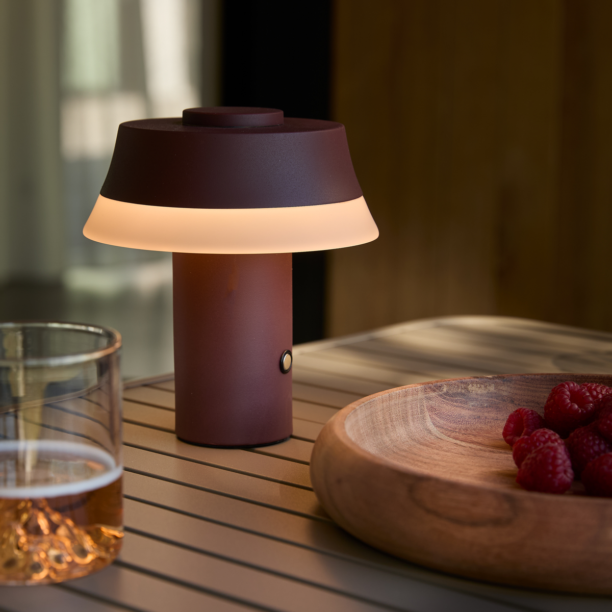 Bærbar vinrød bordlampe, tændt på et udendørs bord ved siden af et glas og en træskål med hindbær.