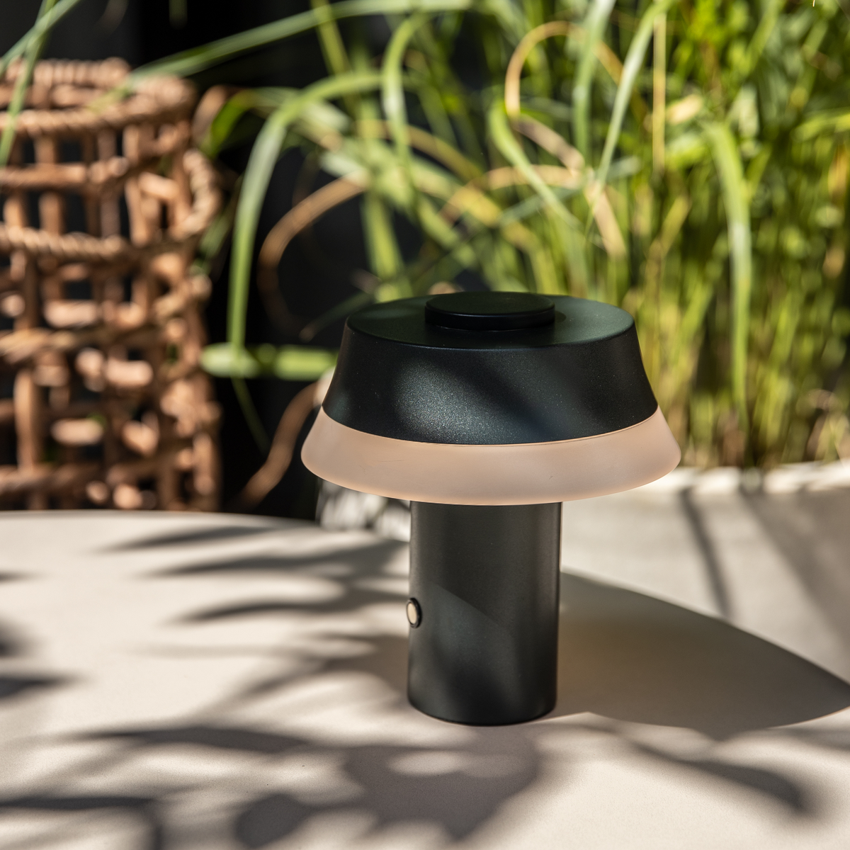 Bærbar sort bordlampe med frostet lysskær på et udendørs bord, belyst af sol med grønne planter i baggrunden.