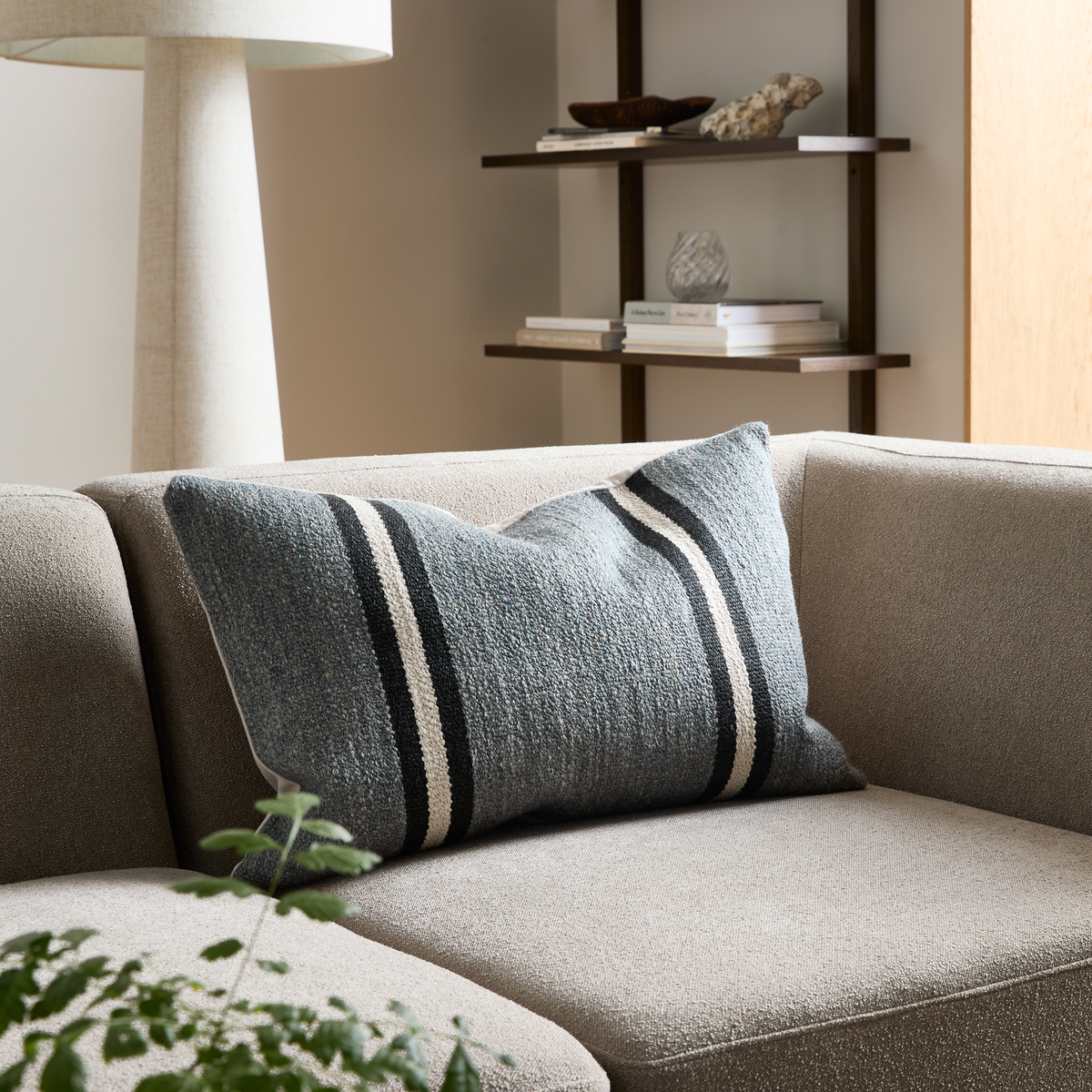 Beige sofa med blågrå stribet aflang pude; gulvlampe, hylder og plante i en minimalistisk stue.