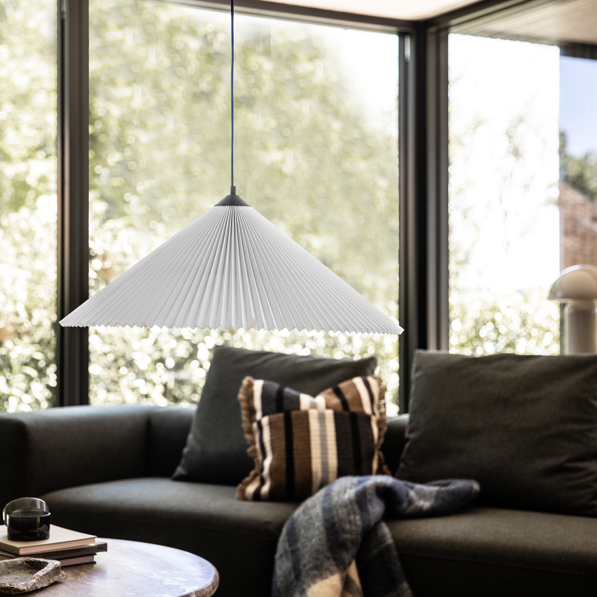 Stue med store vinduer, mørk sofa med stribet pude og plaid, rundt sofabord og en hvid plisseret pendel i fokus.
