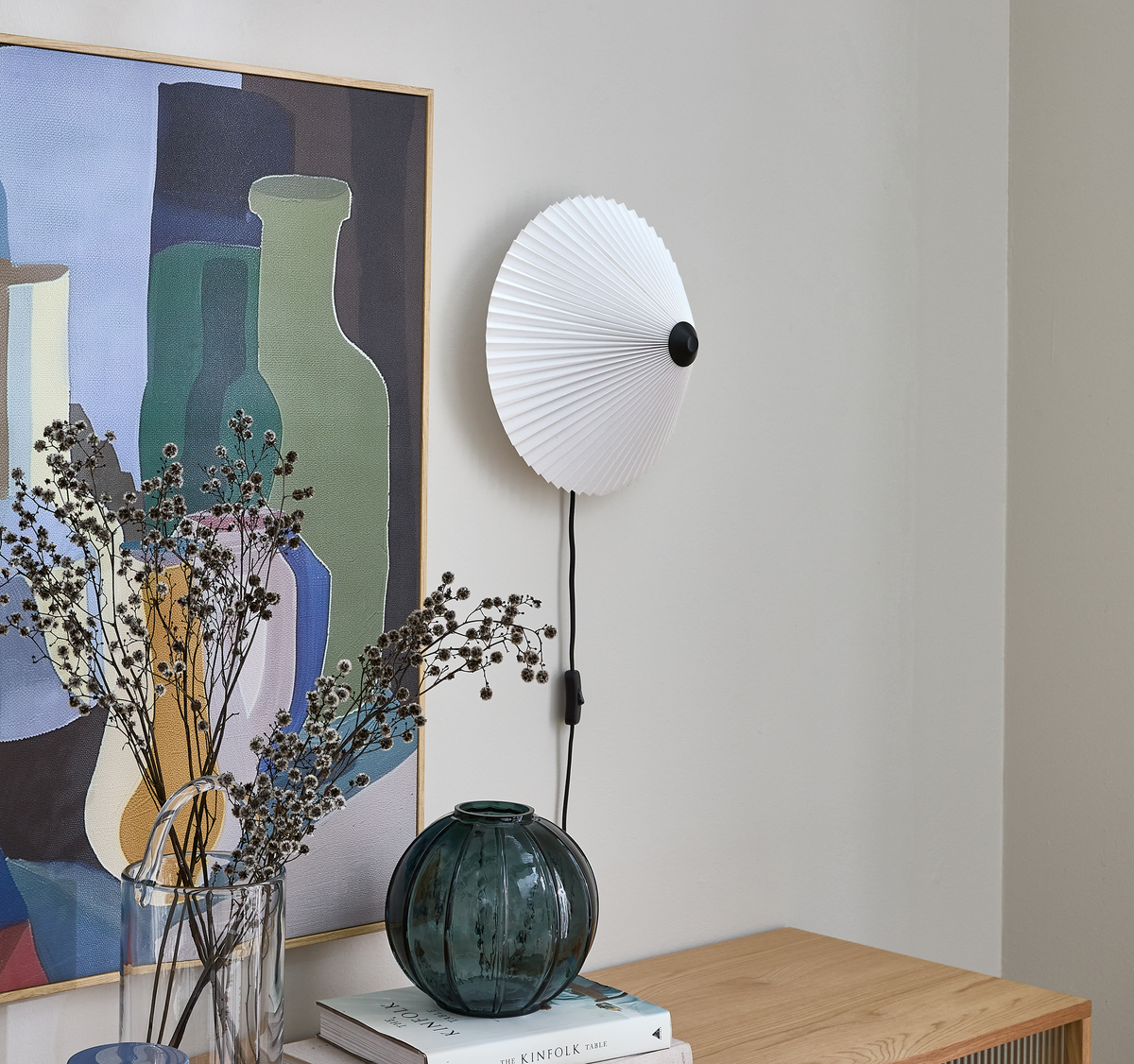 Væglampe med hvid plisseret skærm over en træskænk; abstrakt kunstplakat med flaskemotiver; glasvaser med tørrede kviste og en bogstak med titlen The Kinfolk Table.