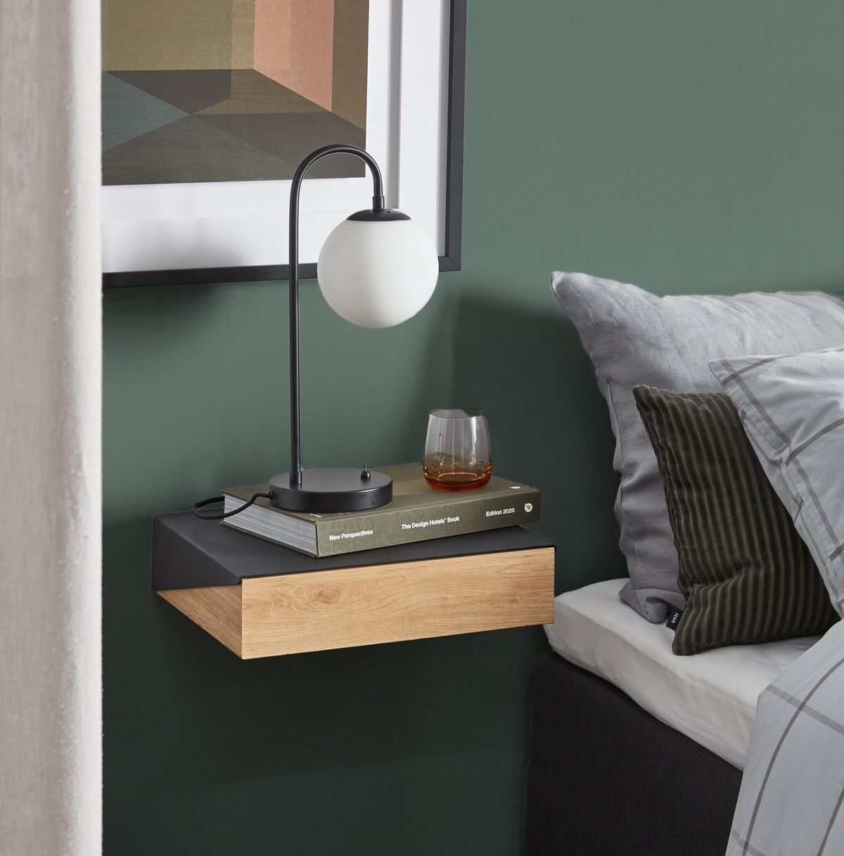 Væghængt natbord i eg og sort med rund kuglelampe, et glas og bogen ”New Perspectives – The Design Hotels Book, Edition 2020” ved siden af en redt seng mod en grøn væg.