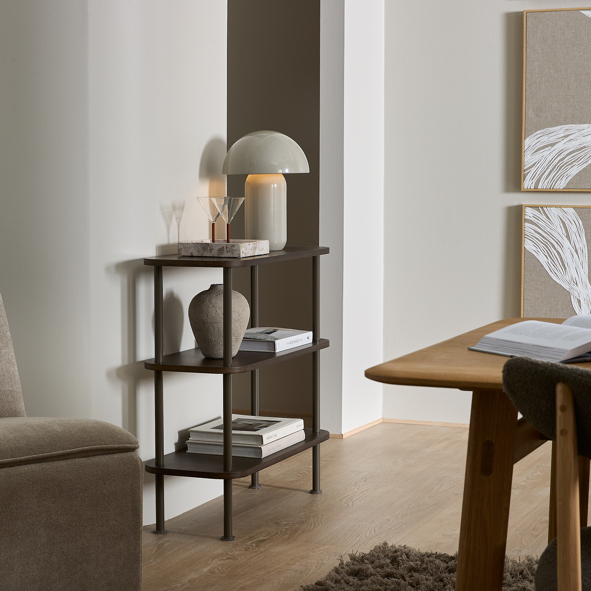 Lille mørkebrun reol med tre hylder i stuen; tændt bordlampe, keramisk vase, to glas på marmorbakke og stakkede bøger ved siden af træbord og vægkunst i neutrale toner.