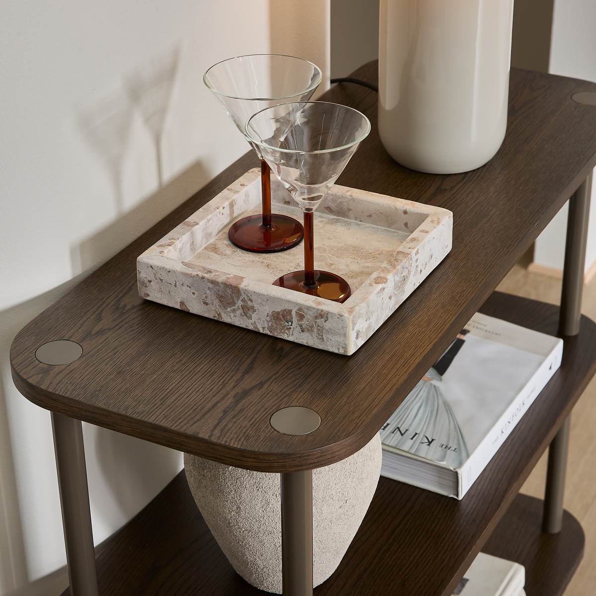 To martiniglas med ravfarvede stænger på et stenfad på en mørk træhylde, ved siden af en keramisk lampe, en vase og en bog med titlen The Kinfolk.