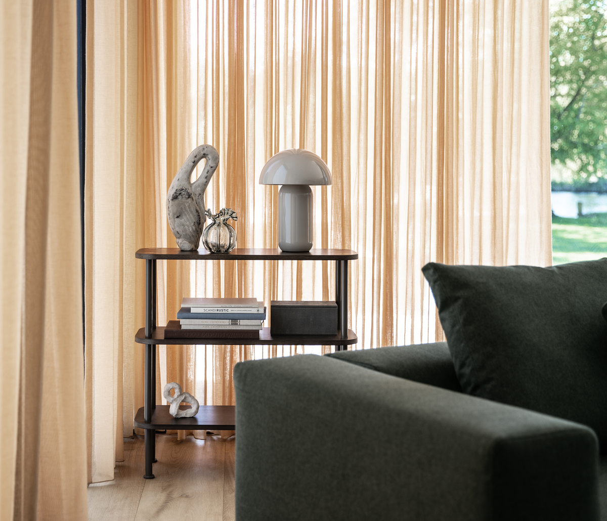 Lille reol i mørkt træ med bordlampe, bøger og skulpturer foran beige gardiner, med en grøn sofa i forgrunden.