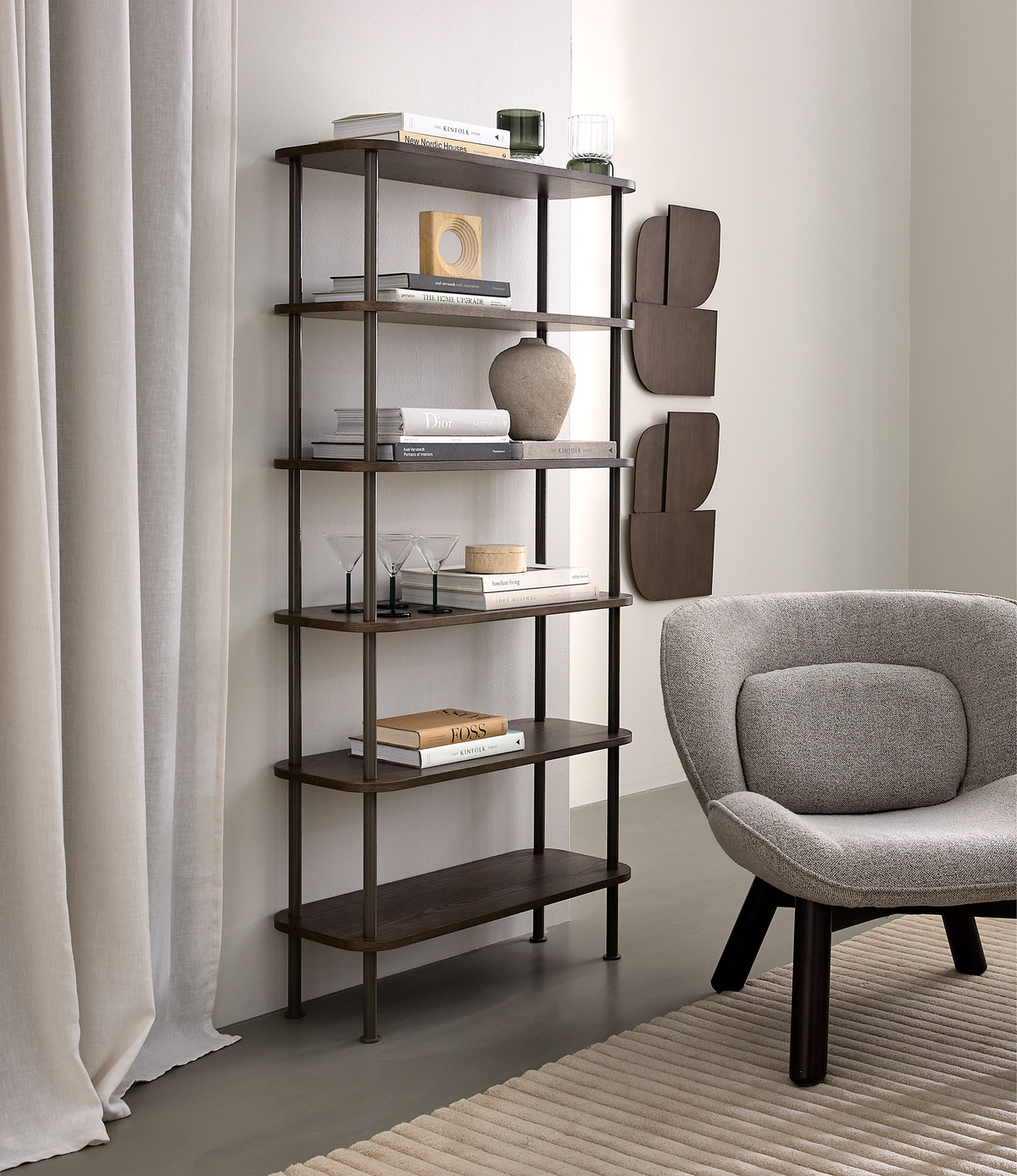 Minimalistisk stue med mørk træreol stylet med bøger (bl.a. Dior og Boss), glas og en keramisk vase; grå polstret lænestol og rillet beige tæppe ved lyse gardiner i dagslys.